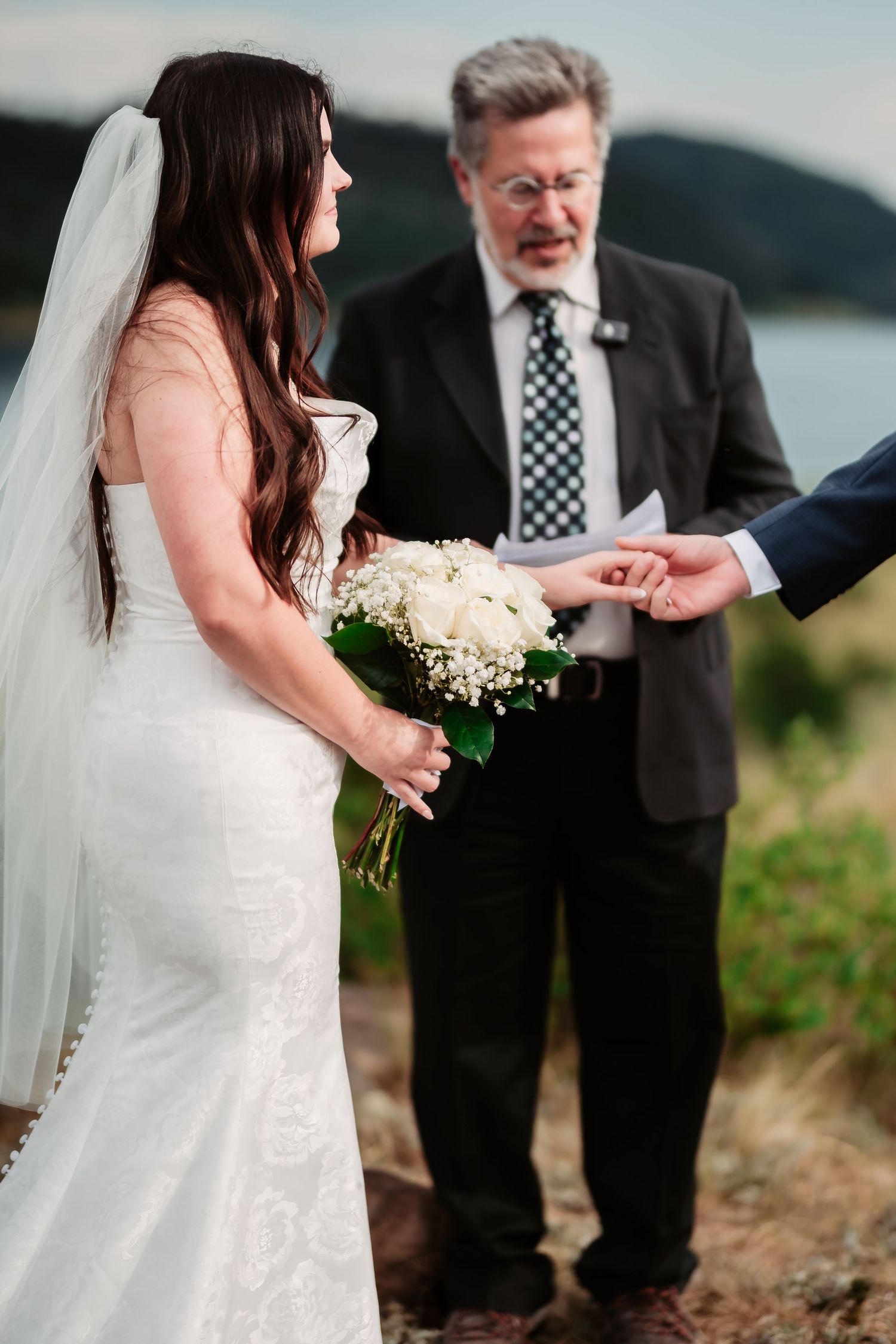 A bride in white dress holds a bouquet during lakeside wedding ceremony with officiant.