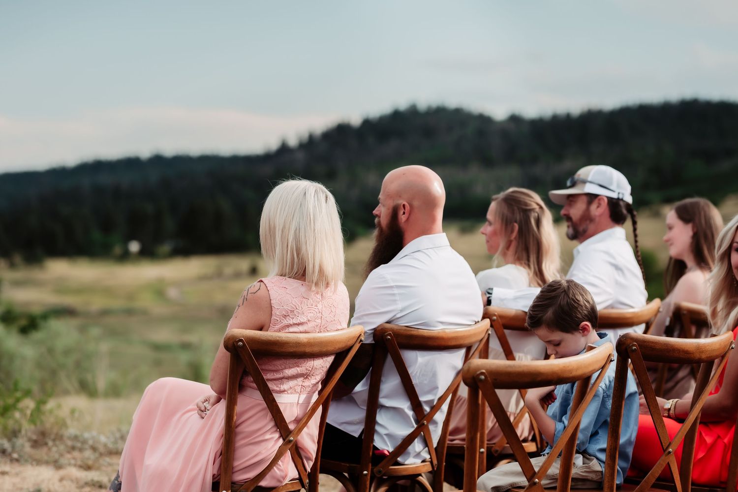 Wedding guests seated in wooden chairs watch the ceremony against a backdrop of mountains and water.