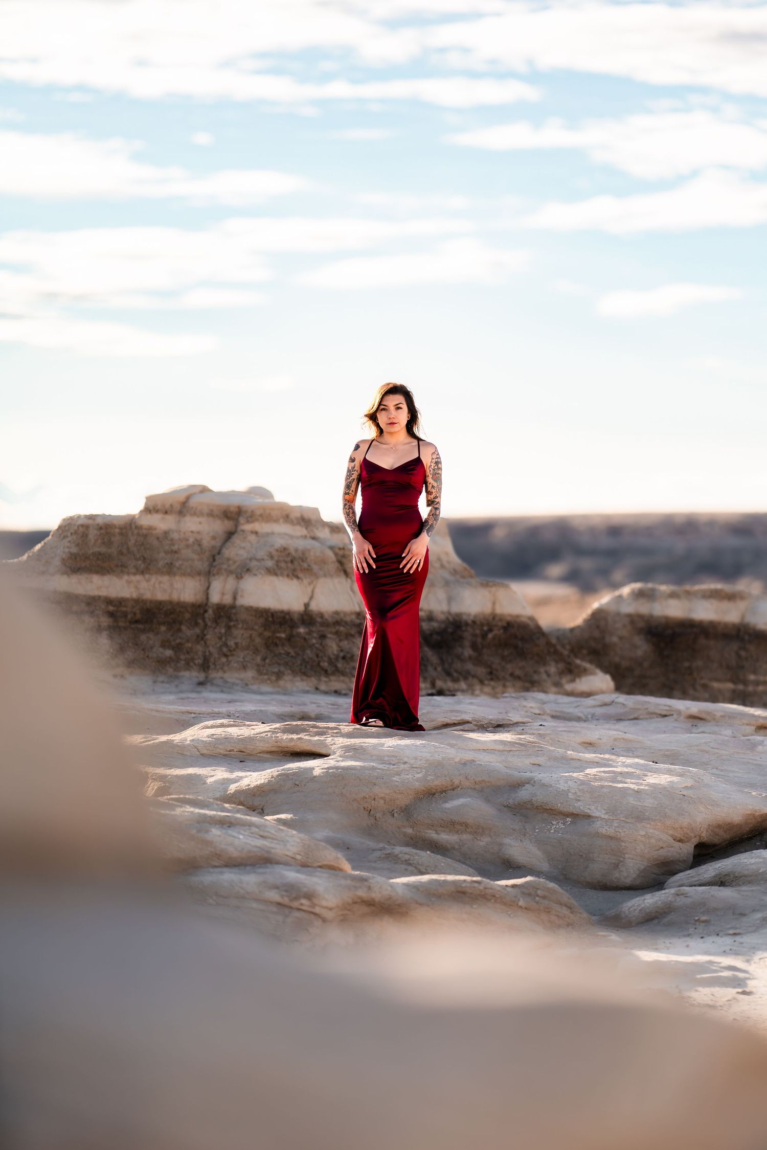 A woman in a flowing burgundy dress poses elegantly on a rocky desert landscape under a blue sky.
