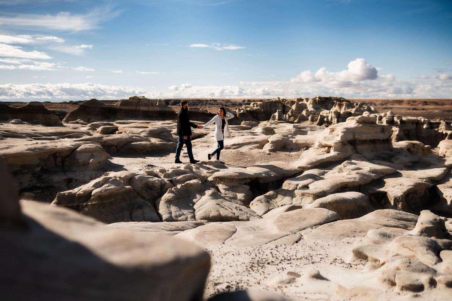 Two silhouetted figures stand on rocky formations in the Bisti Badlands wilderness area under a bright blue sky.