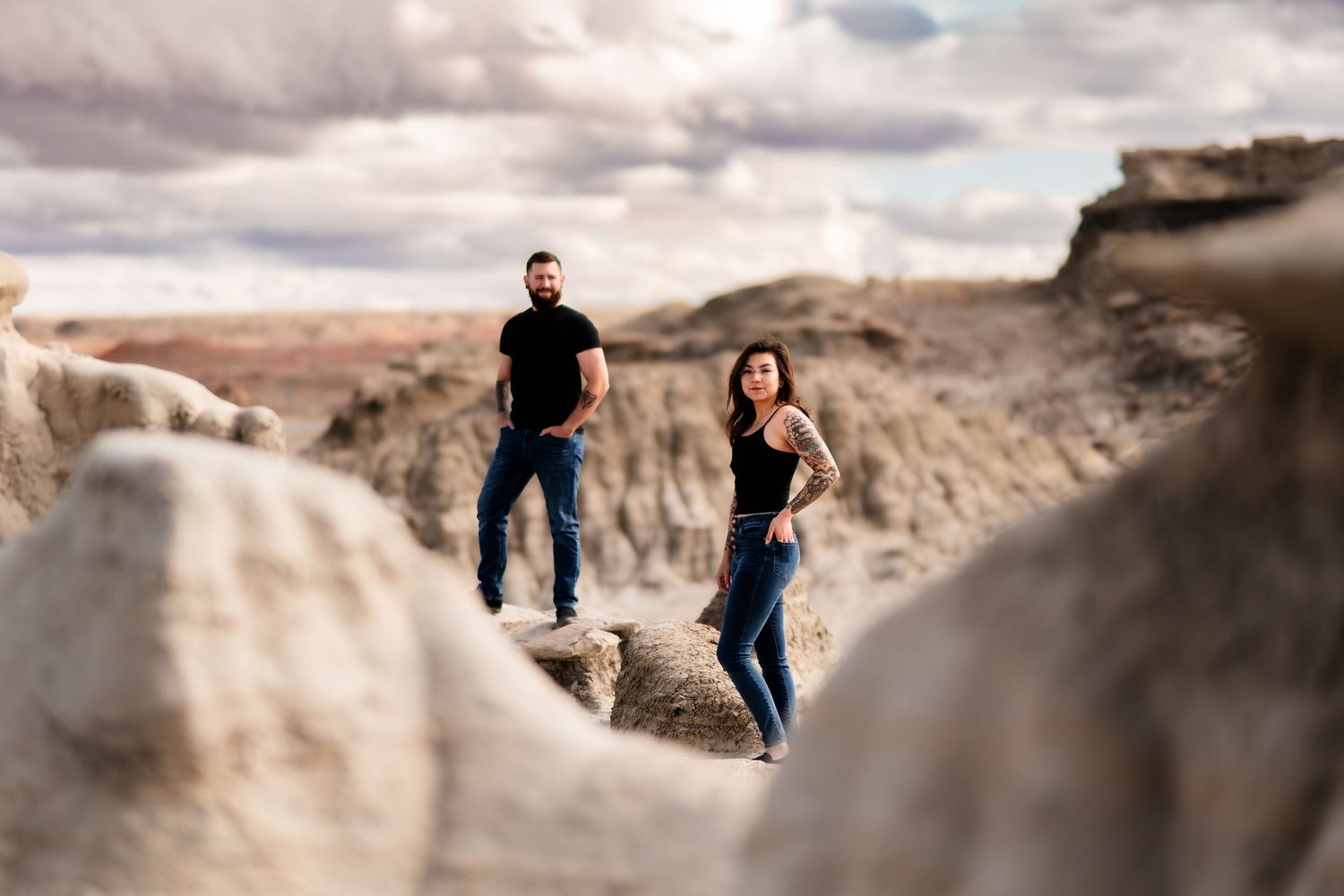 A couple stands among dramatic rock formations in a desert landscape during sunset with moody clouds overhead.