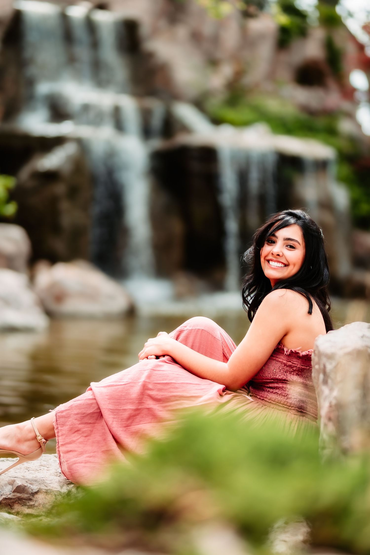 Person in pink dress poses elegantly by a waterfall on rocky terrain with natural scenery.