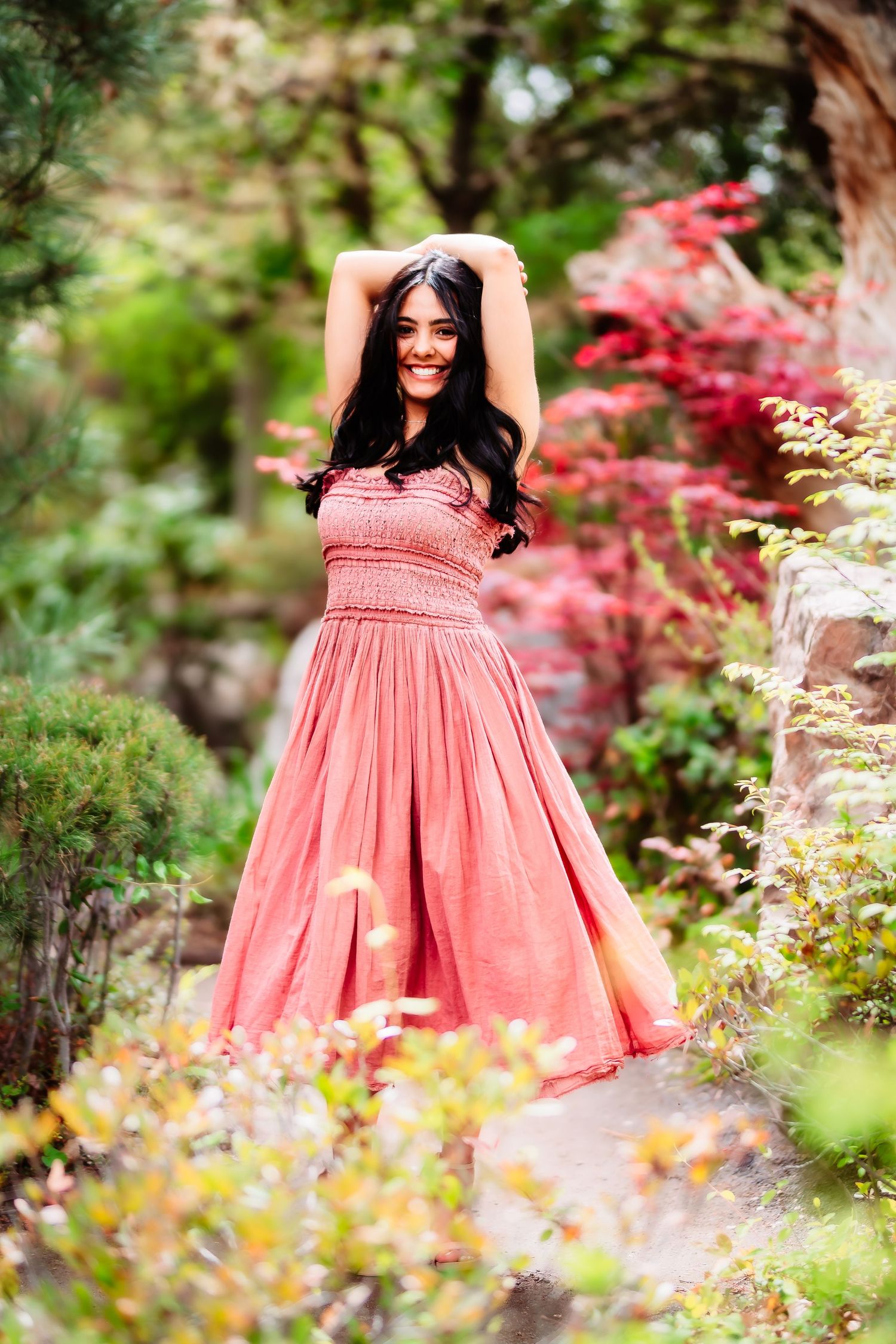 Person in flowing coral dress twirls joyfully in a garden filled with colorful foliage.