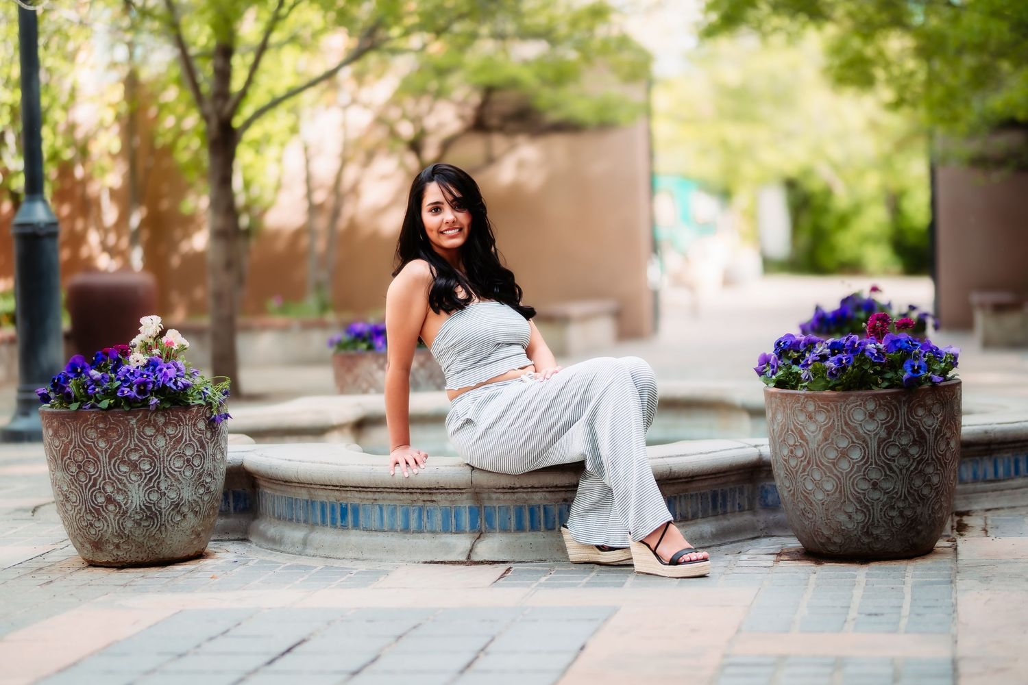 A person in a white dress poses next to decorative flower pots with purple blooms in an outdoor courtyard setting.