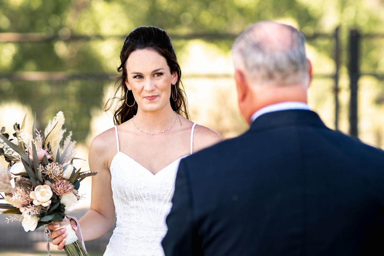 A bride holding a pink and white bouquet during an outdoor wedding ceremony on a sunny day.
