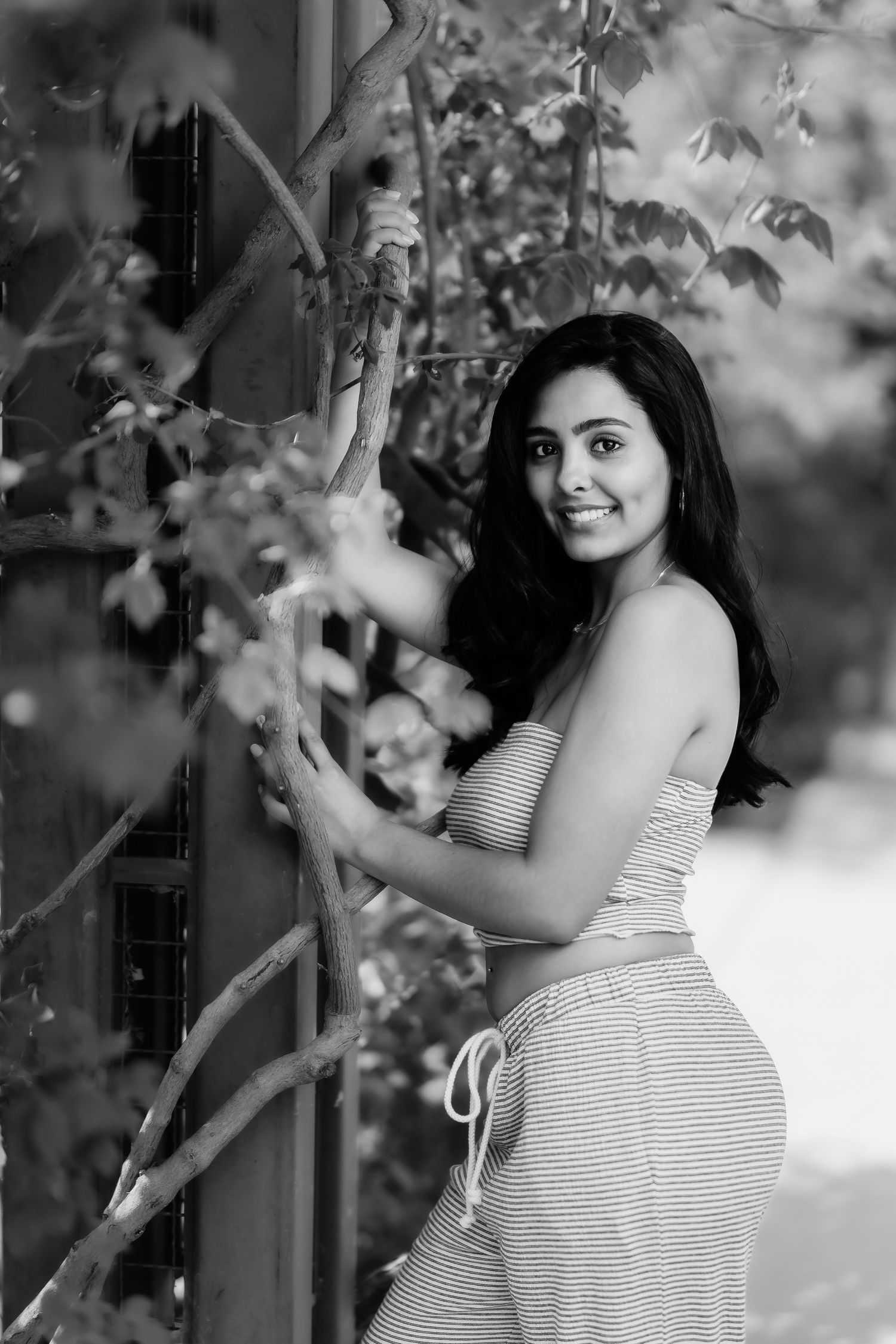 Black and white photo of woman in striped dress posing near foliage and garden trellis.