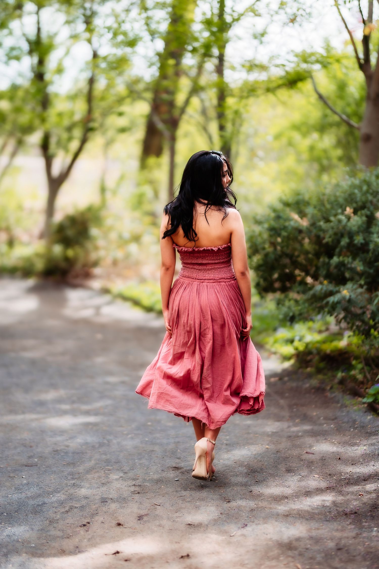 A person in a flowing pink dress walks along a scenic forest path on a sunny day.