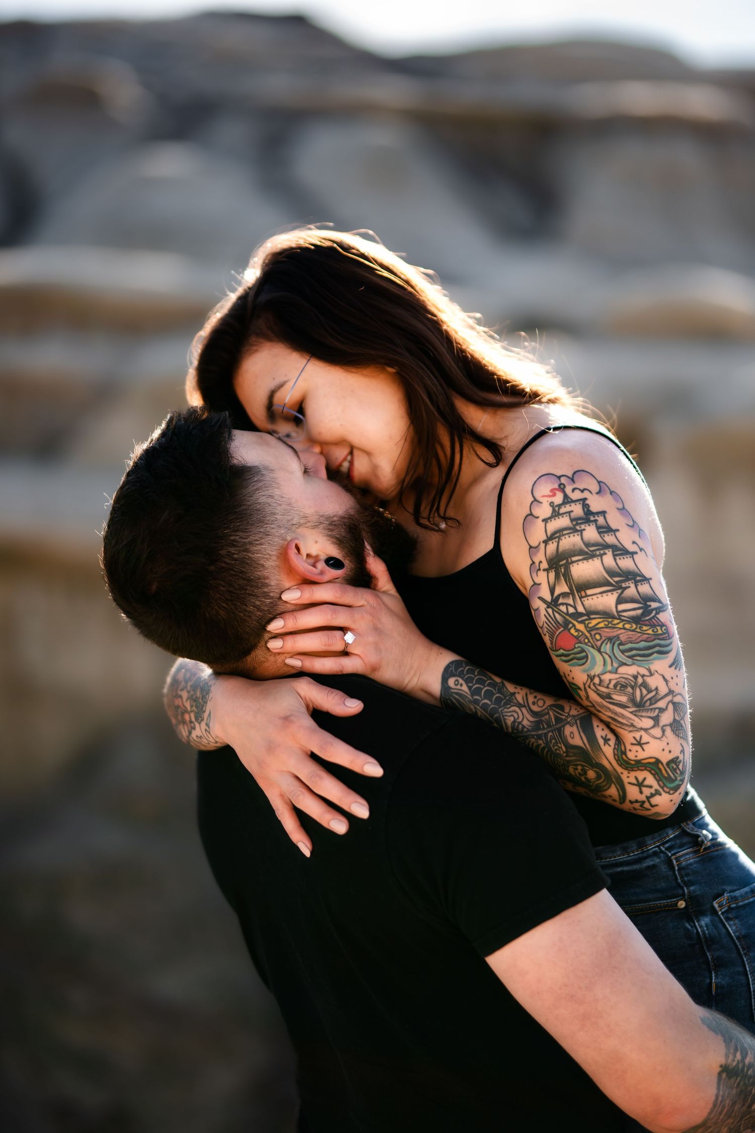 Intimate couple embracing with visible ship sailing tattoo against rocky desert backdrop.