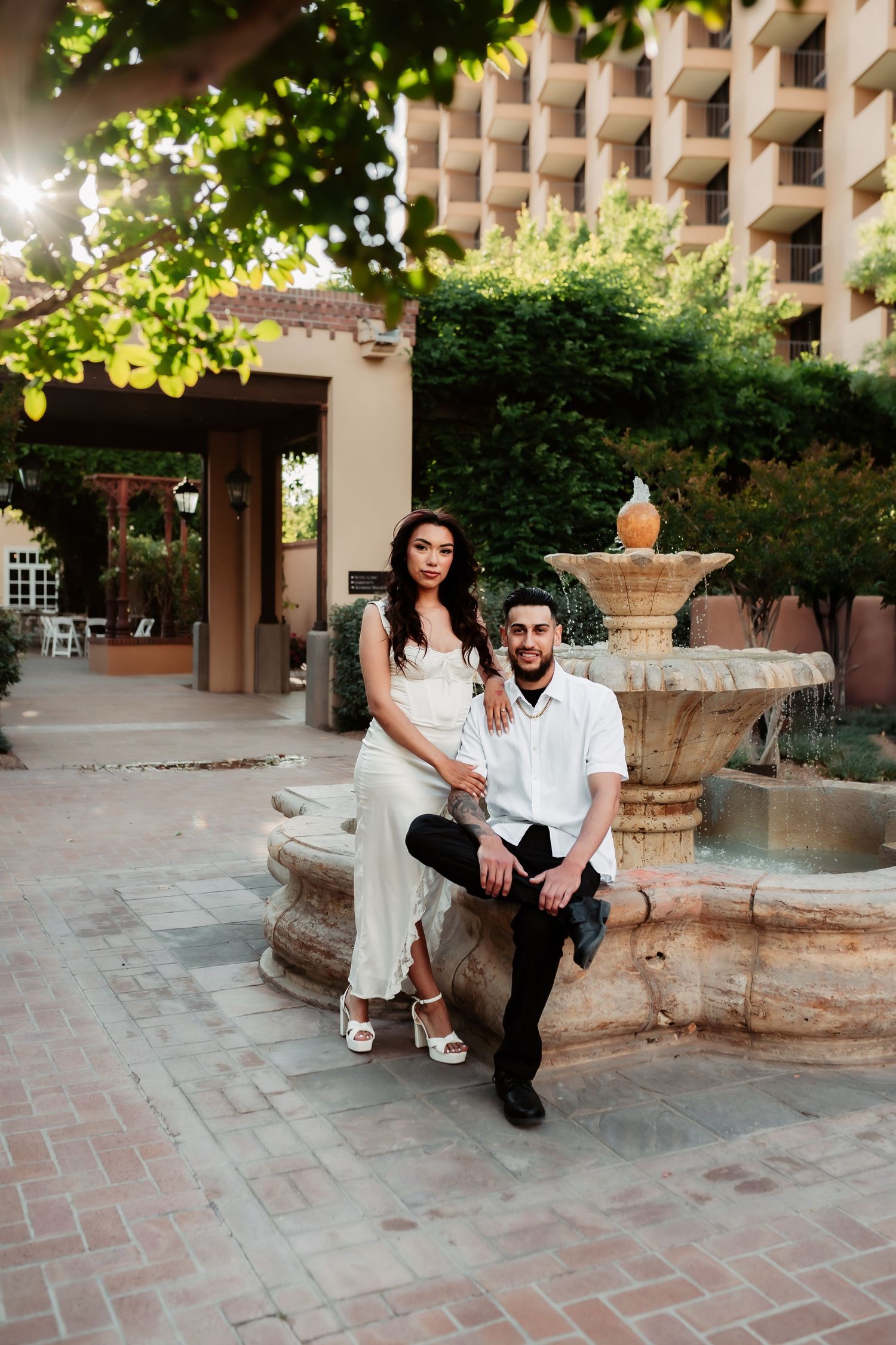 Couple sits casually by an outdoor fountain with modern architecture in the background.