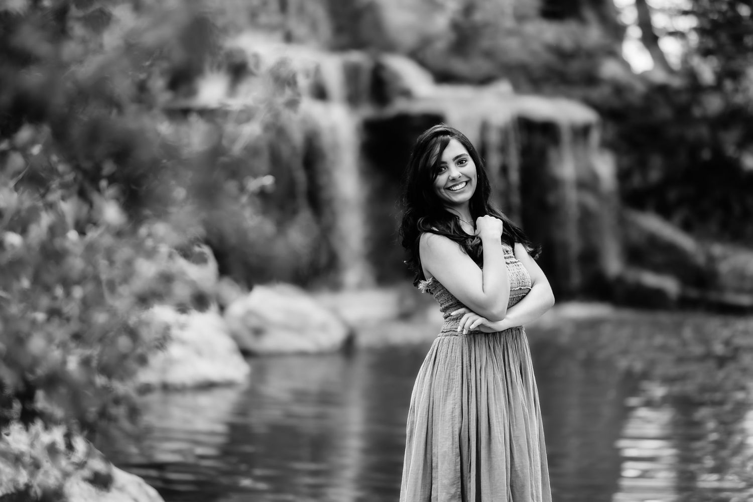 Black and white portrait of someone in a flowing dress standing by a waterfall with arms crossed.
