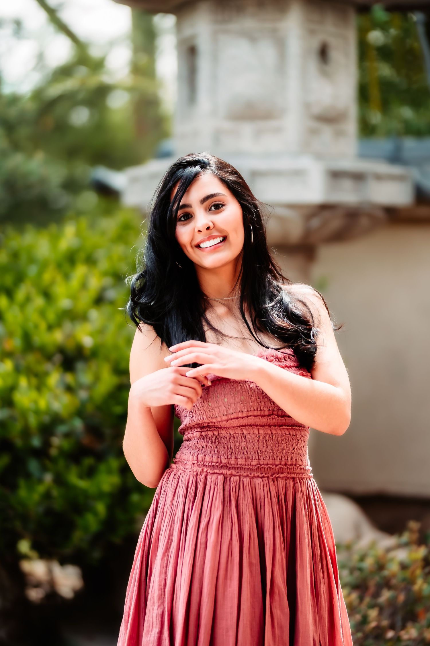 A person in a dusty rose smocked dress poses outdoors with a bright smile against a garden background.
