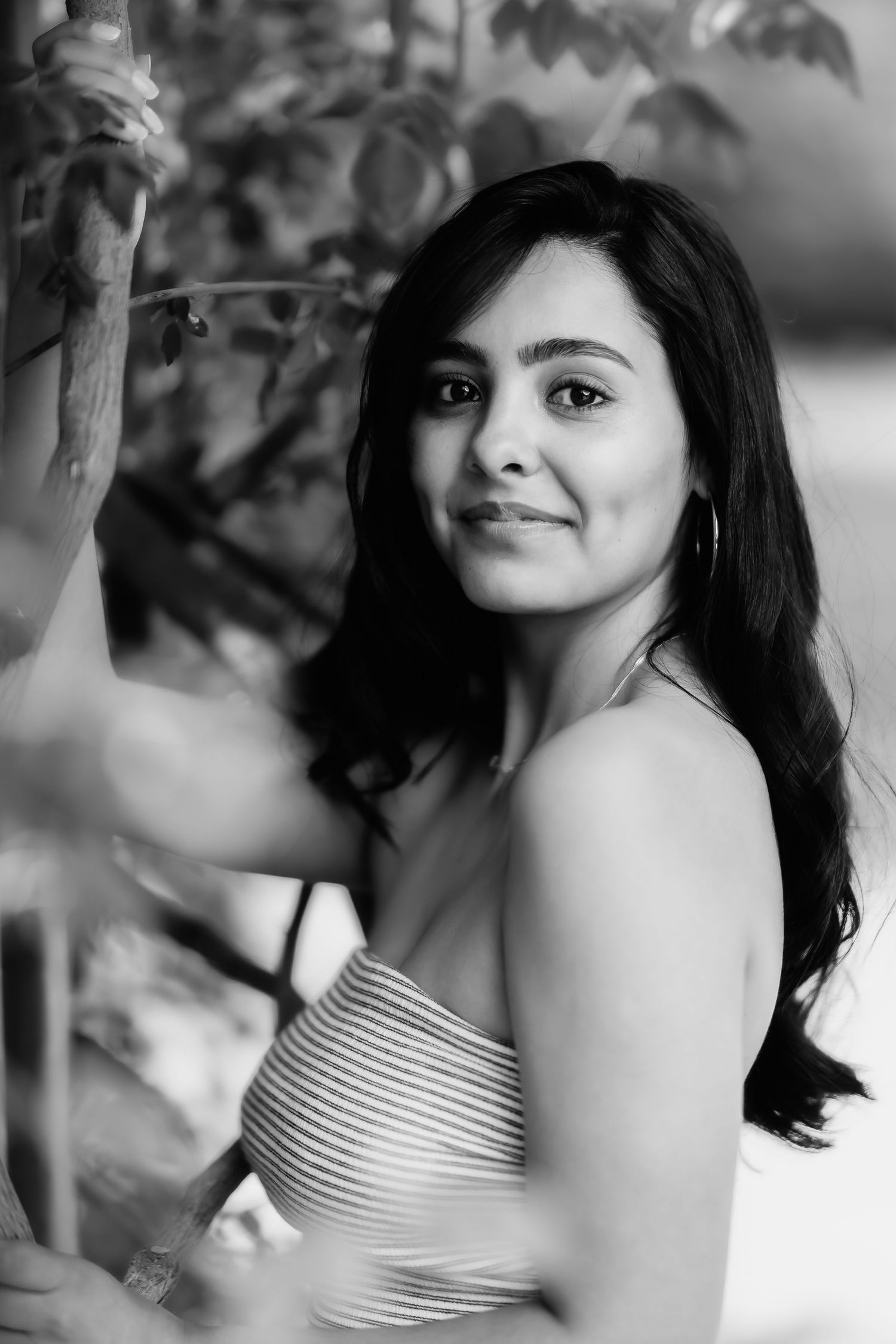 Black and white portrait of a person with long dark hair in a striped top, posing outdoors among foliage.