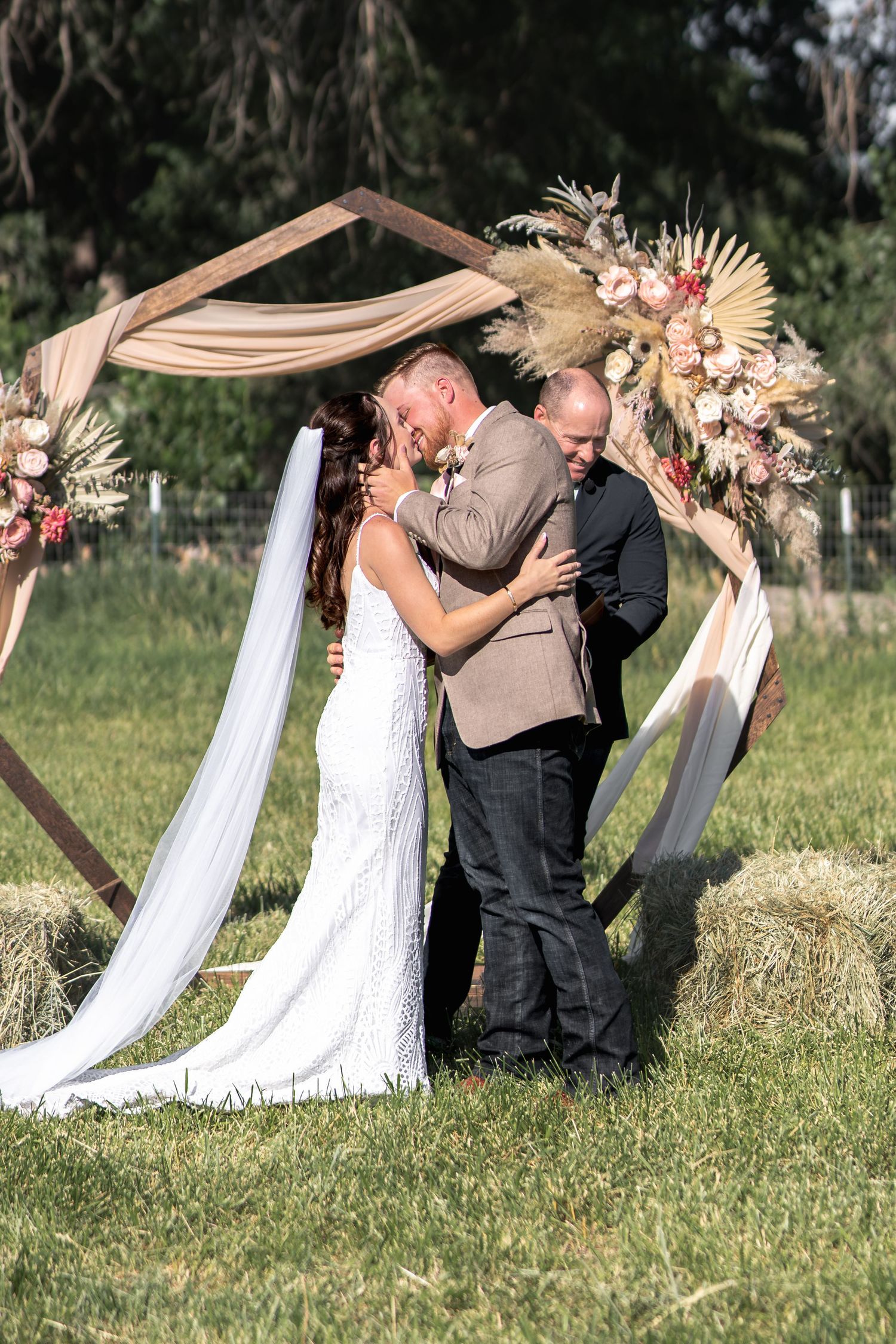 Outdoor wedding ceremony with couple embracing under a floral arch with pink and white draping.