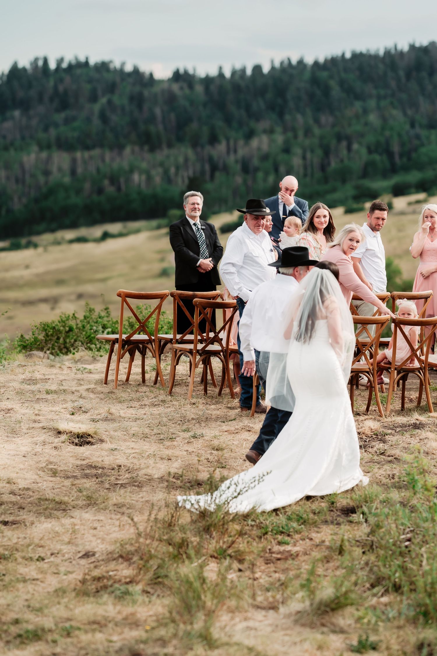 Wooden cross-back chairs set up for an outdoor mountain wedding ceremony with evergreen forest backdrop.