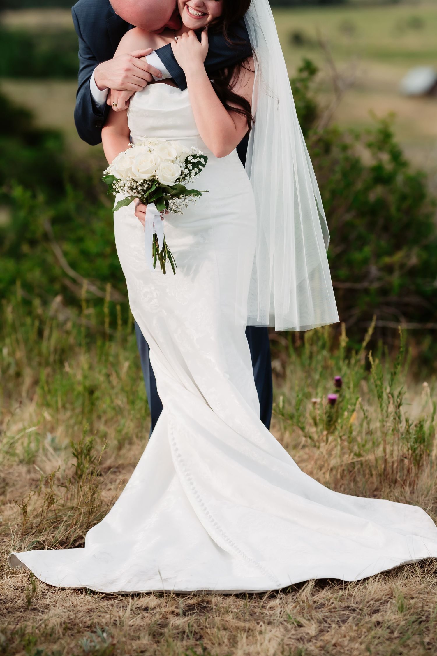 Close-up of an elegant white wedding dress with trailing train and small floral bouquet.