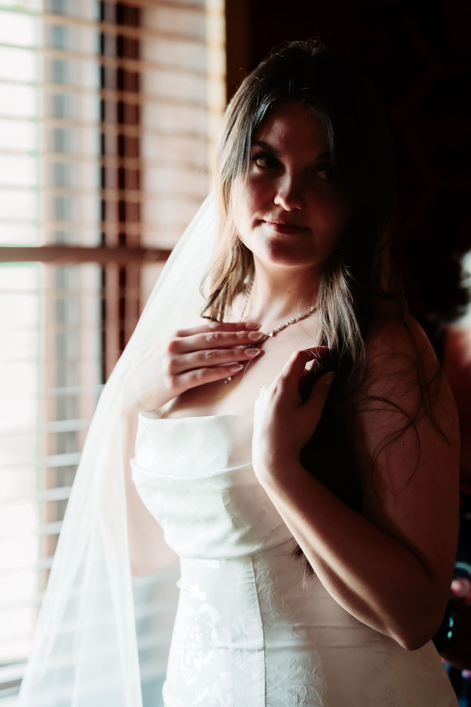 A delicate white wedding dress with veil creates an elegant silhouette by a window with venetian blinds.