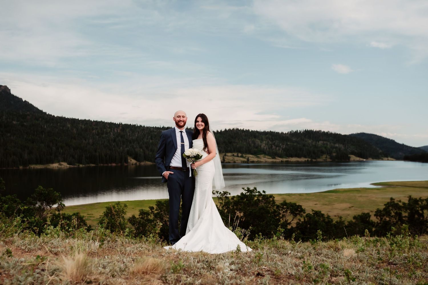 Newlyweds pose for portraits on a hillside overlooking a scenic lake and mountains.