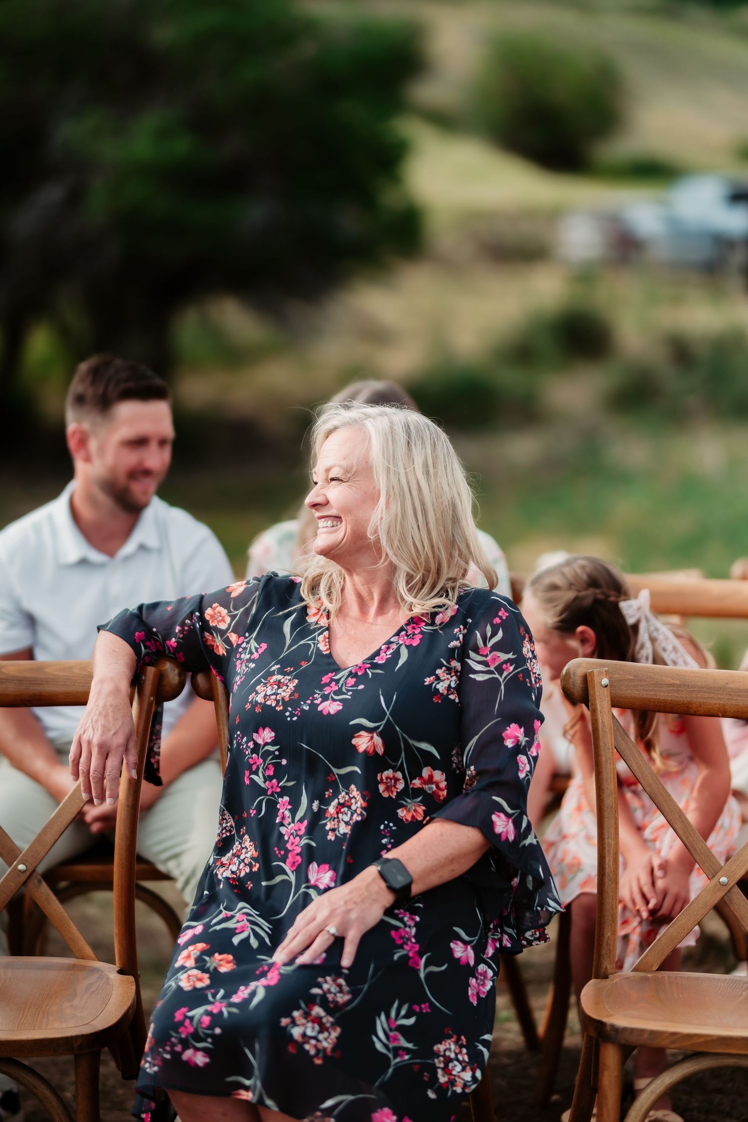 Guest wearing floral dress laughing joyfully at outdoor event with wooden chairs.