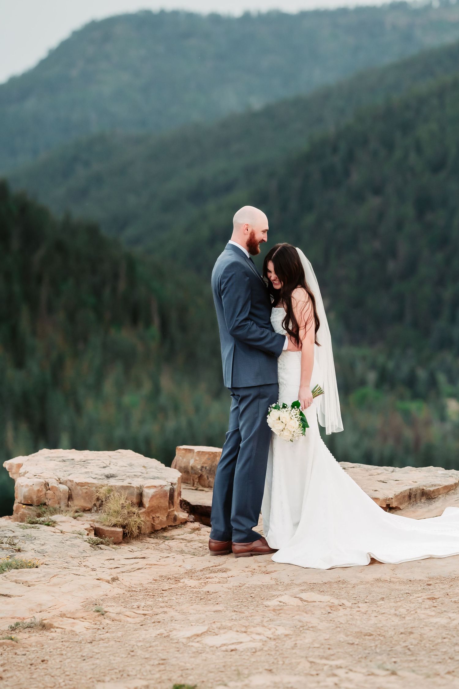 A wedding couple stands on a rocky mountain overlook sharing an intimate moment against dramatic scenery.