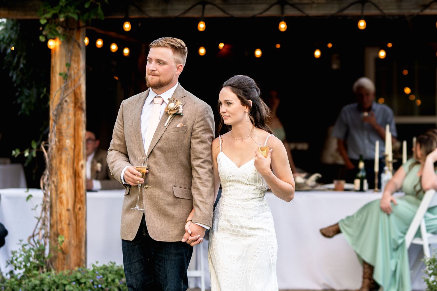 Newlyweds walking hand in hand under string lights at outdoor reception.