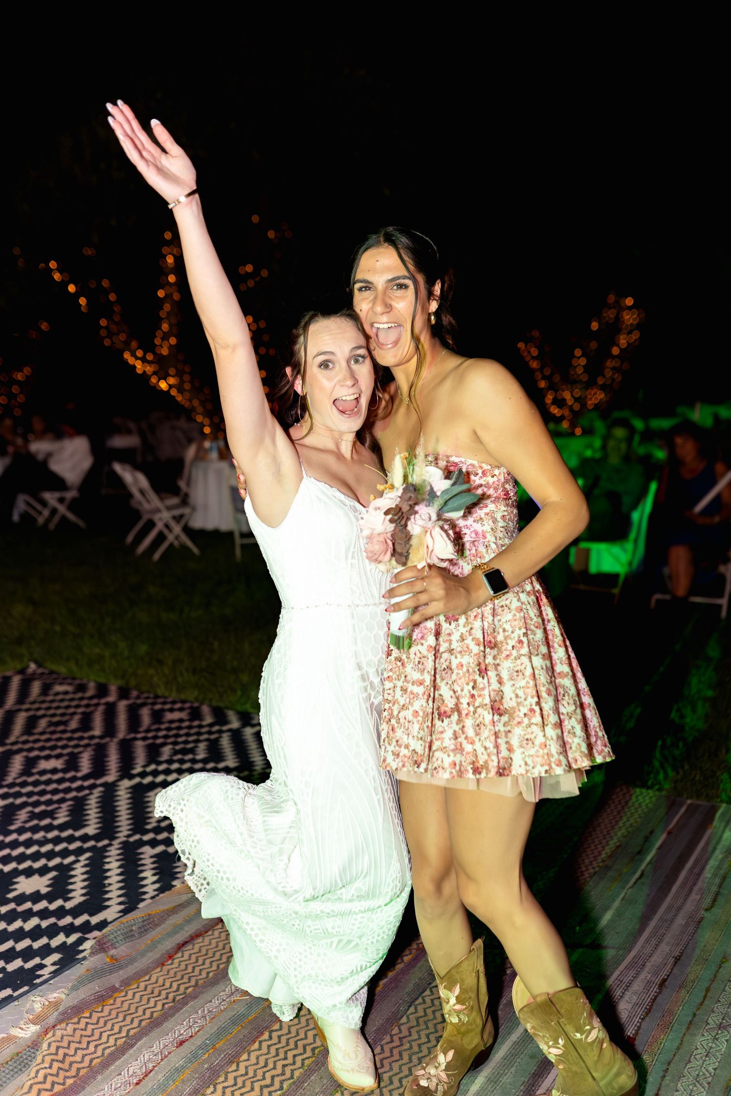 Wedding celebration with bride and friend dancing outdoors at night with festive lighting.