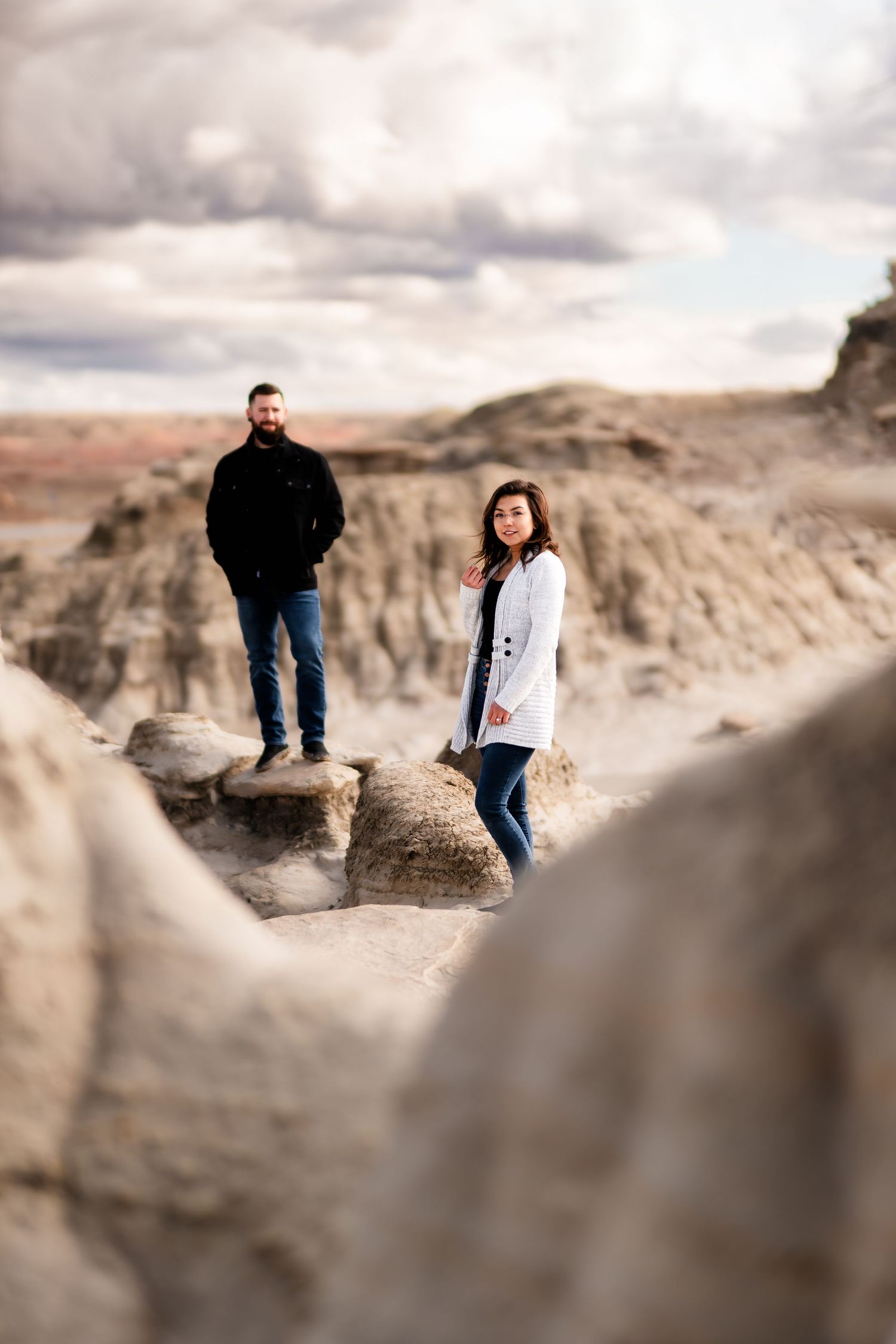 Couple stands on dramatic rock formations in a desert badlands landscape under moody clouds.
