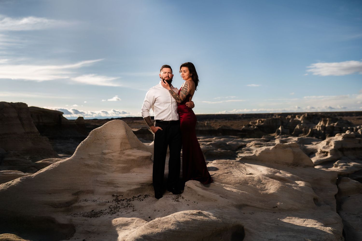 A couple poses together on rocky terrain during sunset in a desert landscape.