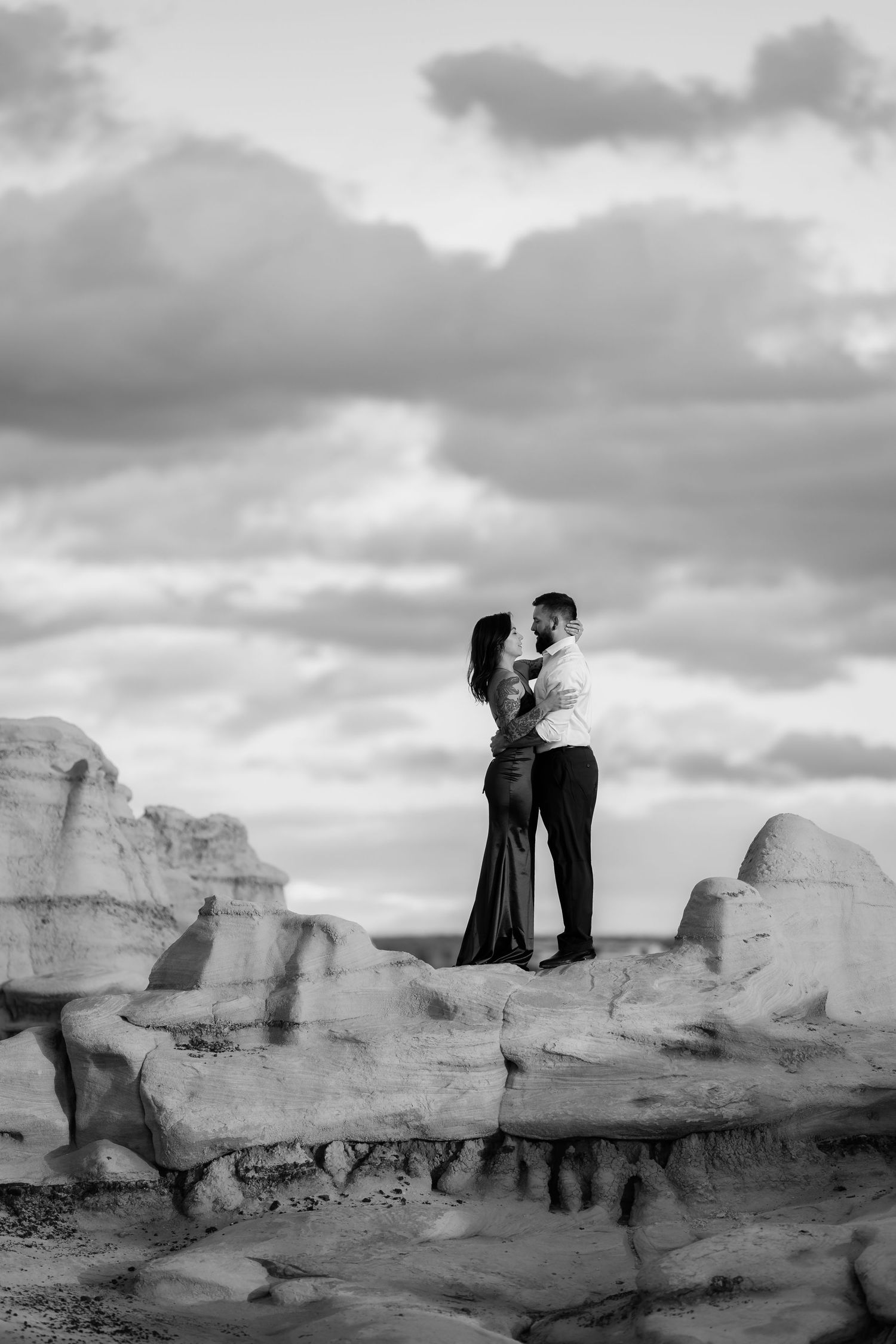 A romantic couple embraces on dramatic rocky terrain under moody cloudy skies in black and white.