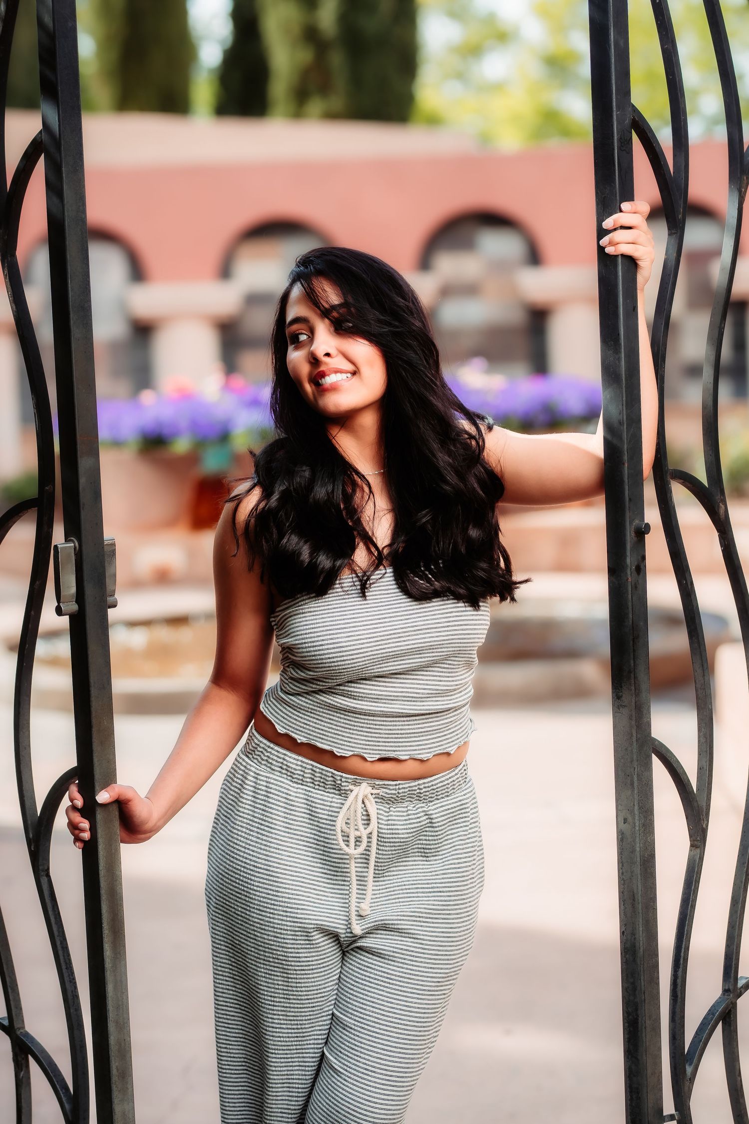 A woman in casual grey loungewear poses between decorative metal gates with a pink building in the background.