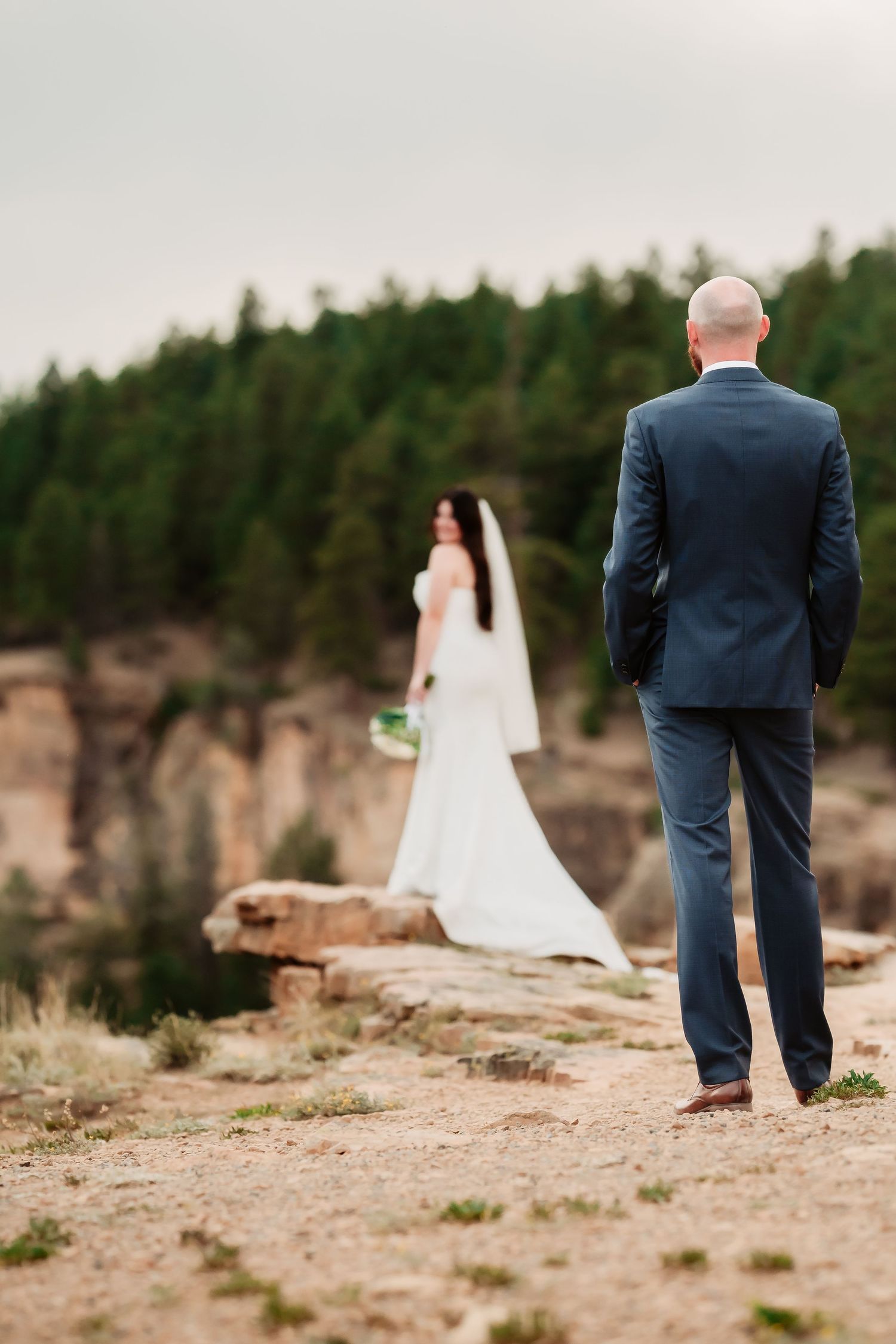 A groom in a navy suit watches his bride from a distance on a rocky mountain ledge.