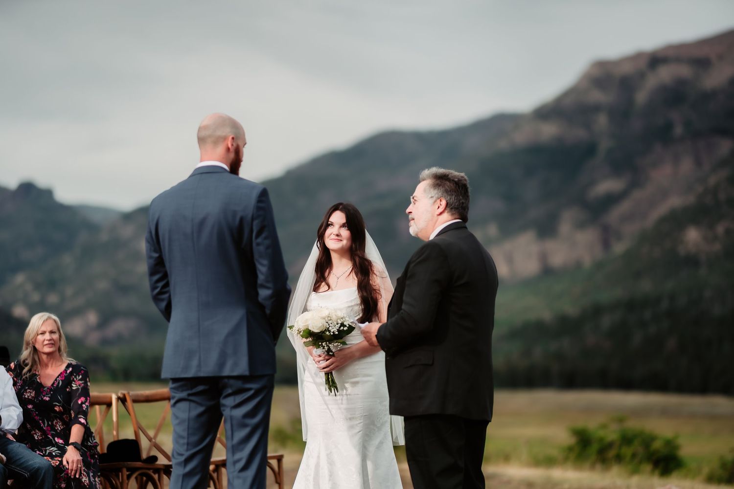 A wedding ceremony takes place against a scenic mountain backdrop as guests observe from wooden chairs.