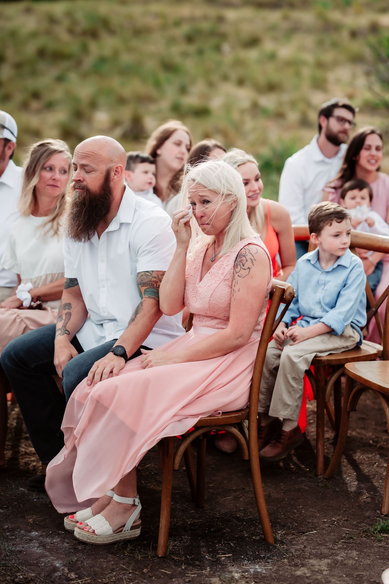 Wedding guests are seated on wooden chairs outdoors showing emotional reactions during a ceremony.