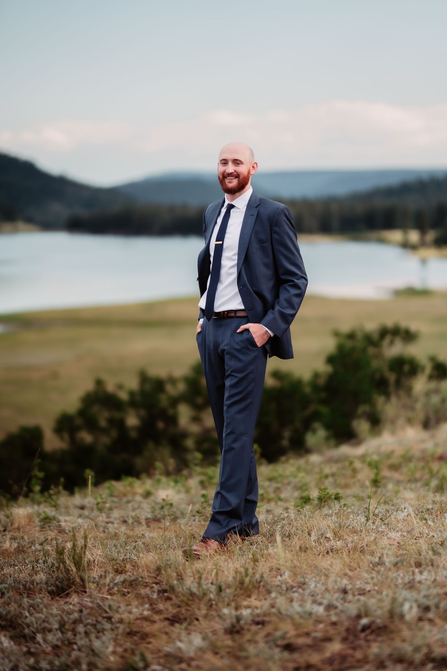 Groom in navy suit poses on hillside with lake and mountains in background.