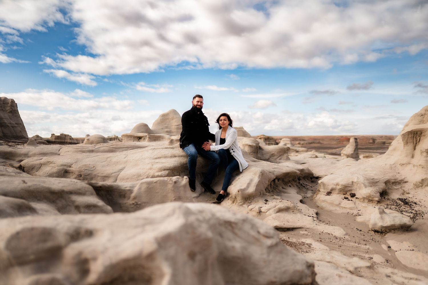 A couple sits together on unique rock formations in a desert landscape under puffy white clouds and blue sky.