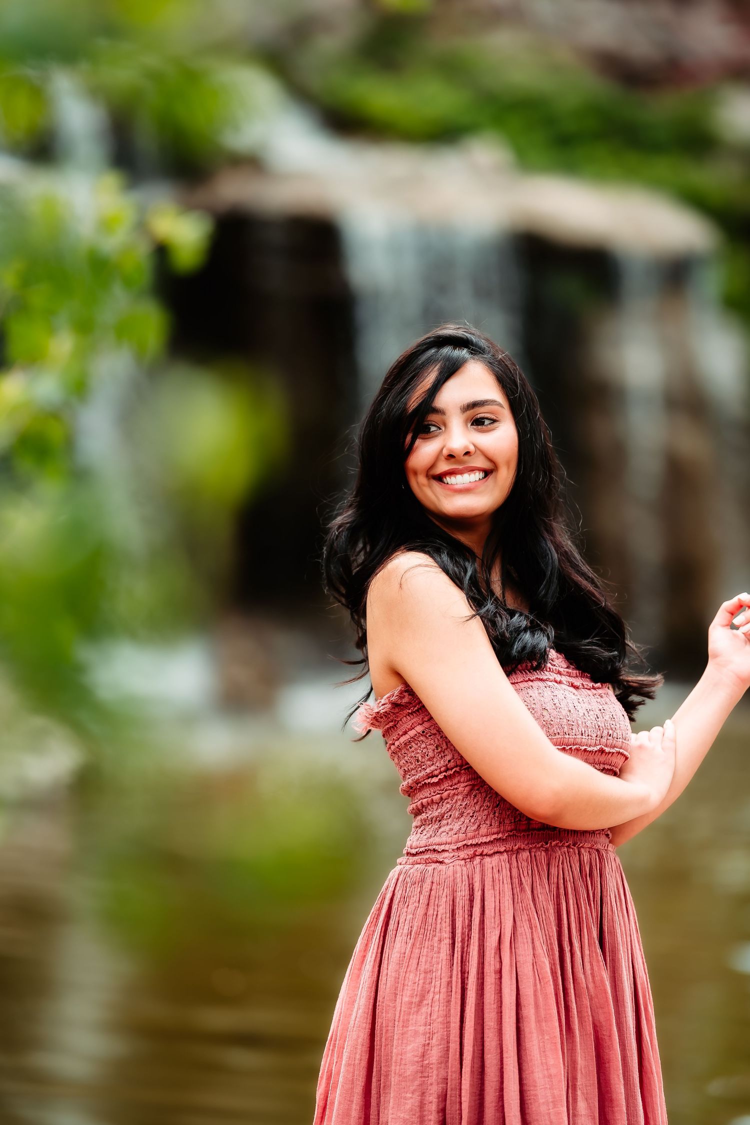 Portrait of someone in a dusty rose dress smiling and posing outdoors near a waterfall.