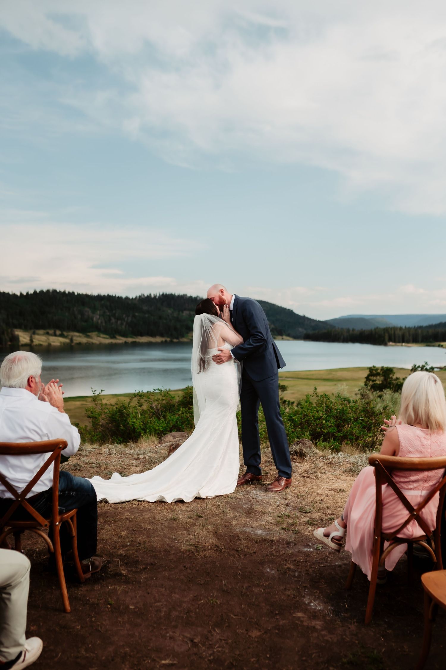 Newlyweds share a moment during mountain lakeside ceremony with guests seated in wooden chairs.