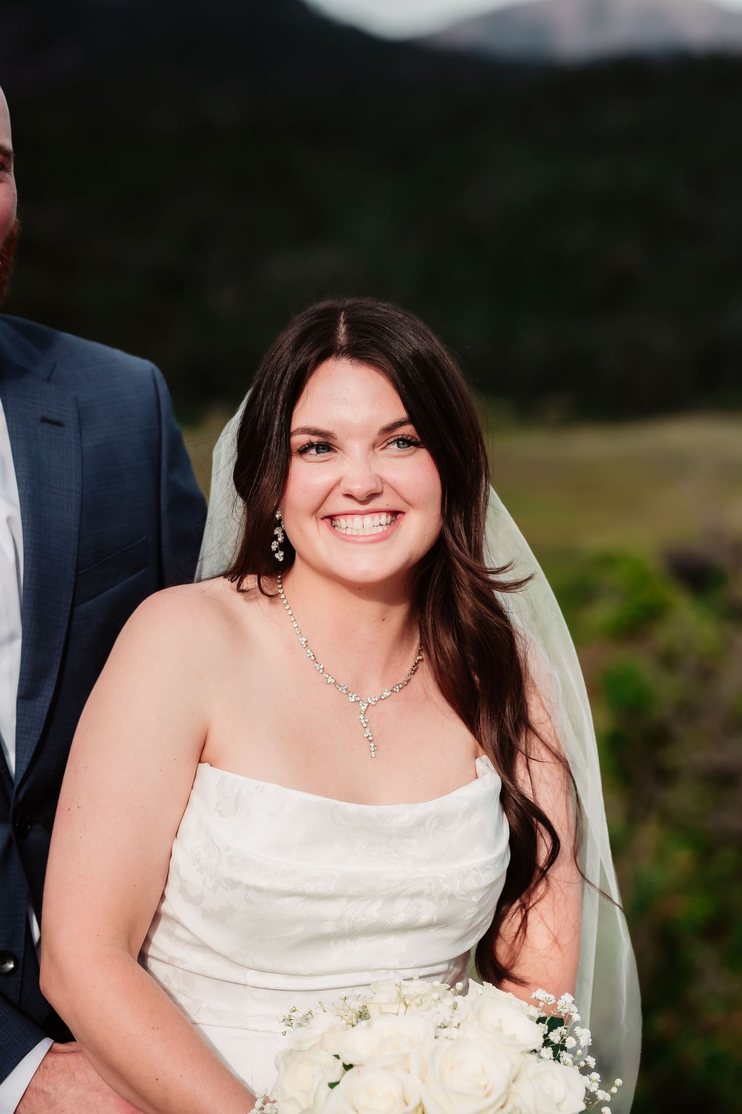 A radiant bride in a white dress holds a bouquet of white roses during an outdoor ceremony.