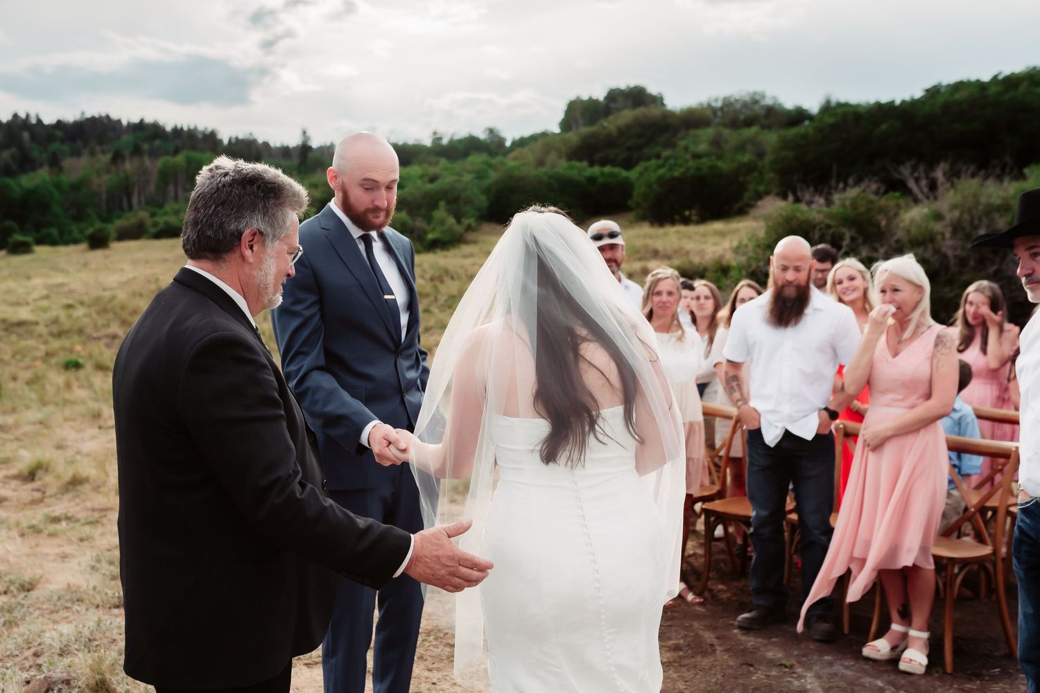 An intimate outdoor wedding ceremony takes place with guests seated on wooden chairs in a mountain setting.