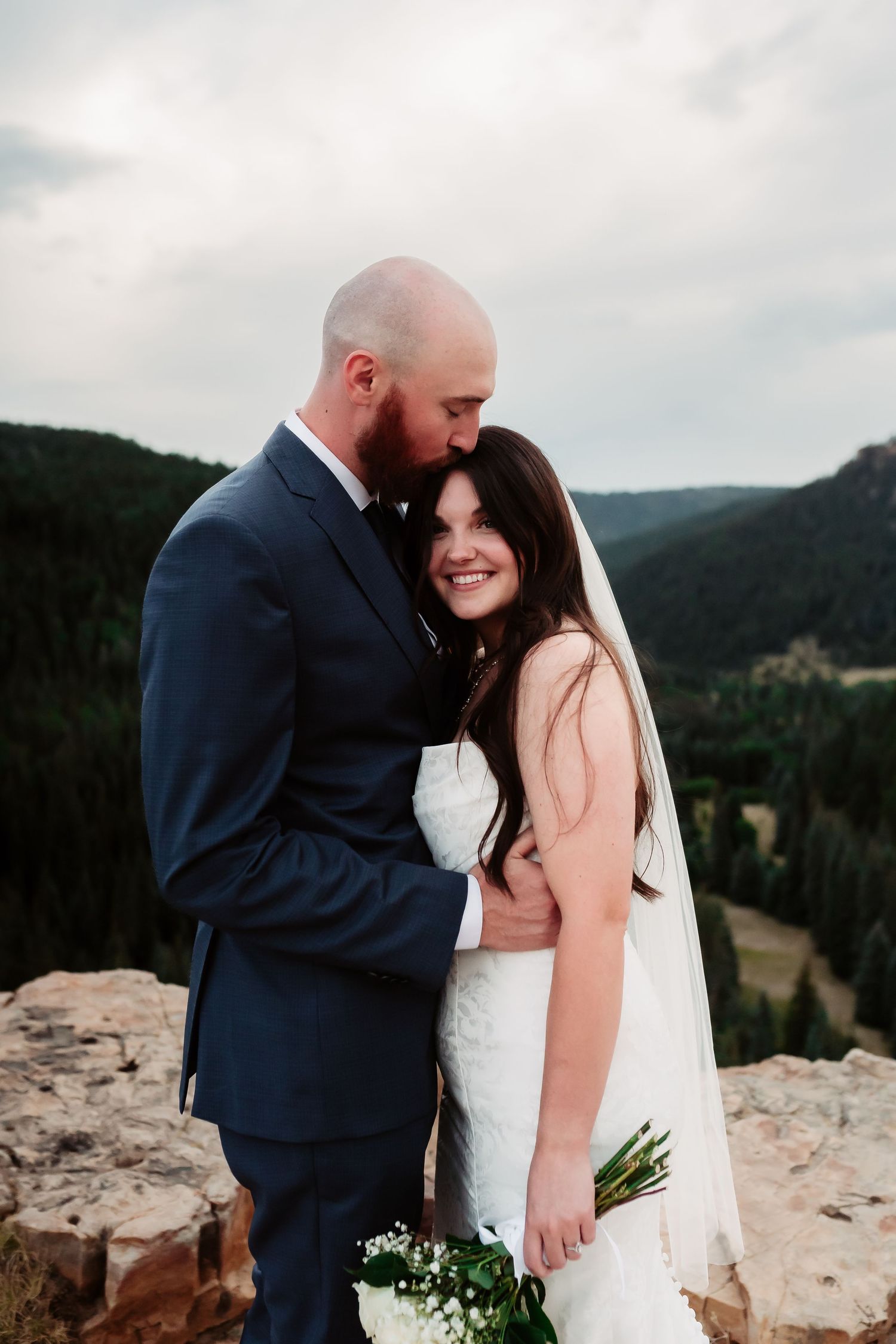 Wedding couple embraces on rocky mountain ledge with scenic valley backdrop.