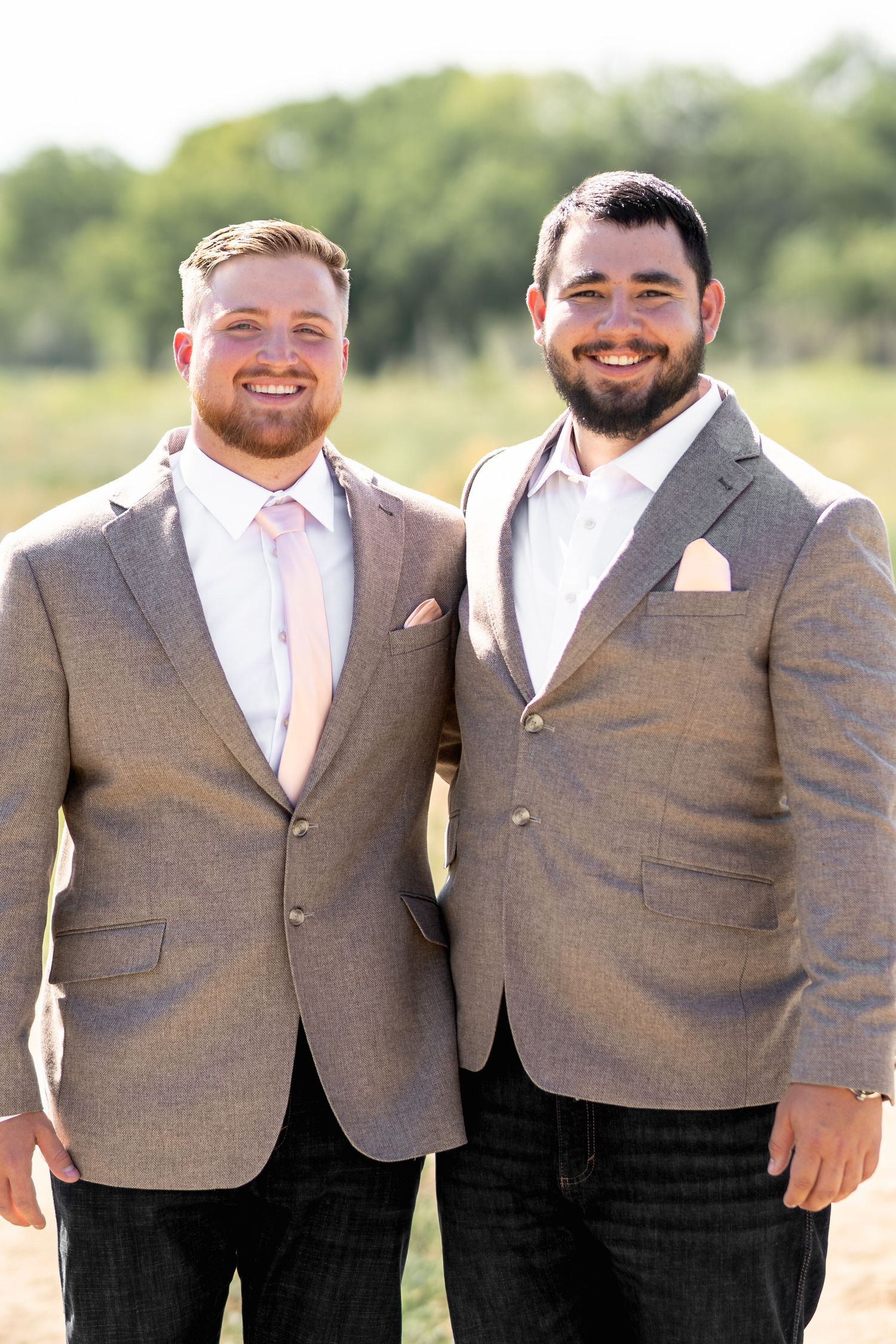 Two groomsmen in matching gray suits and pink ties smile together outdoors.