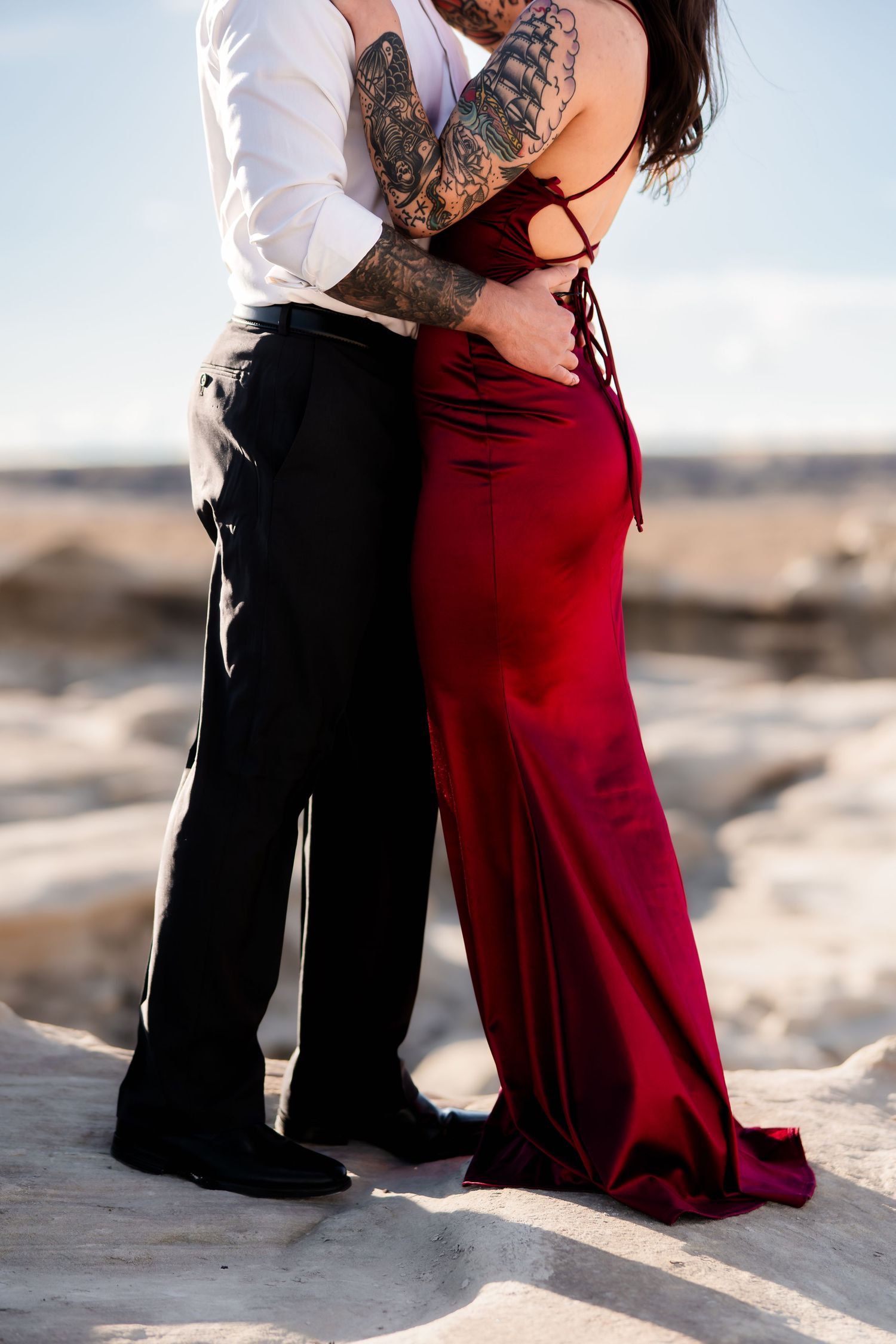 A couple embraces on a rocky surface, with one wearing a flowing red velvet dress and the other in formal attire.