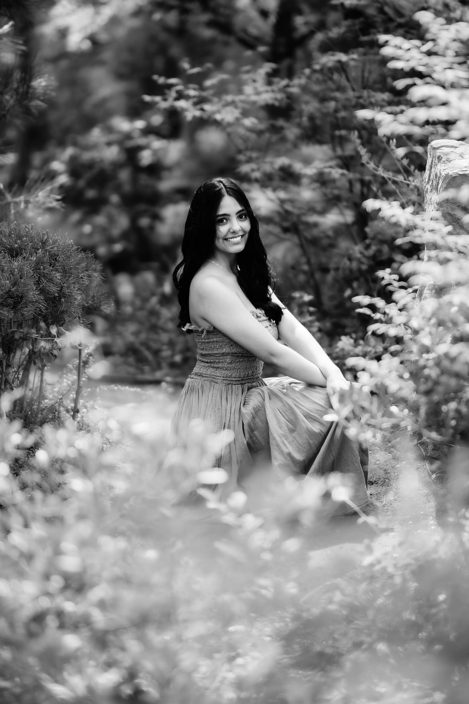 Artistic black and white photo of someone in flowing dress surrounded by garden flowers.