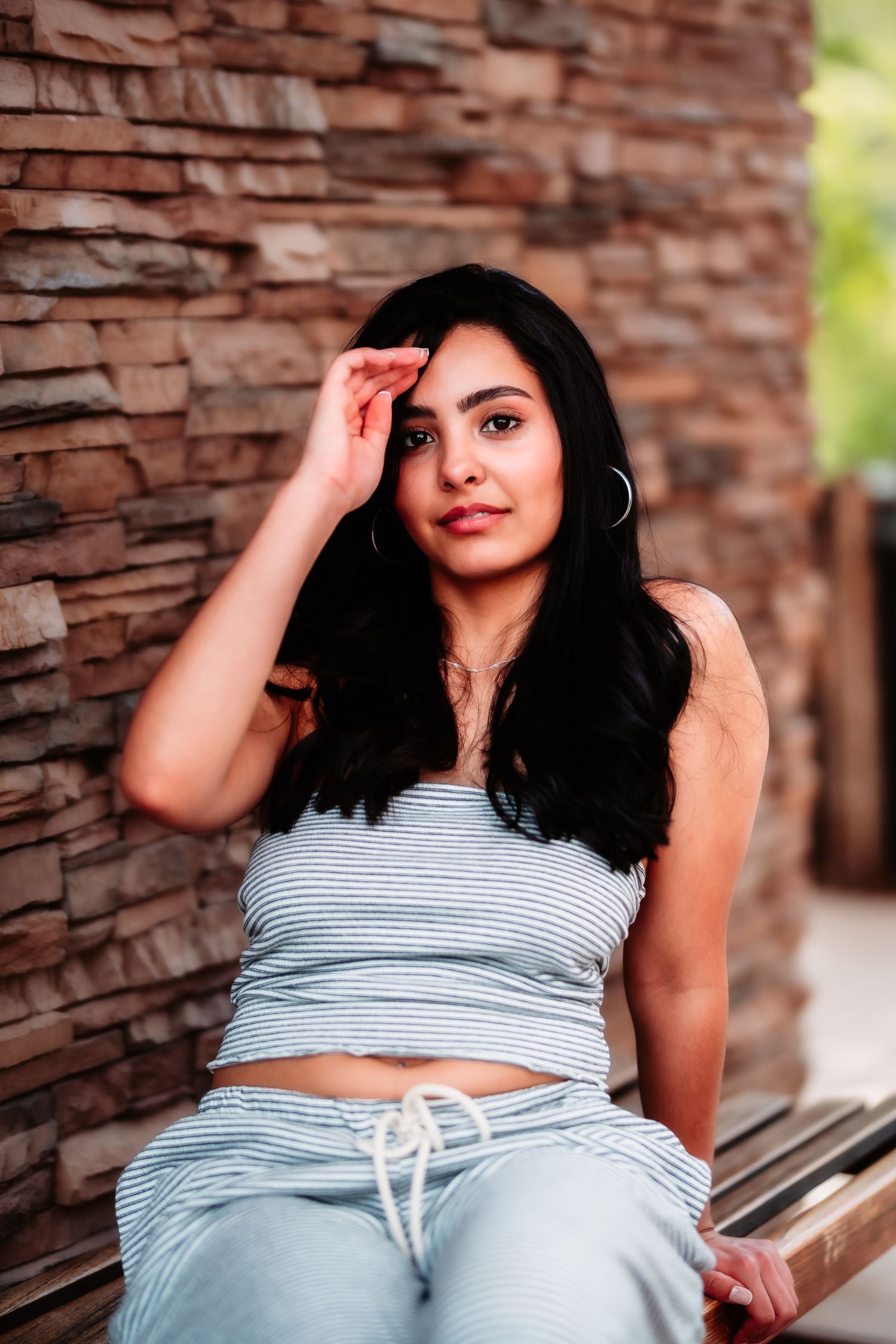 Person in striped crop top and drawstring pants poses casually against rustic brick wall in outdoor portrait.