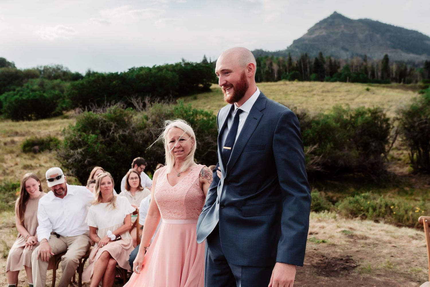 Wedding party dressed in pink bridesmaid dresses and suits poses outdoors against mountain backdrop.