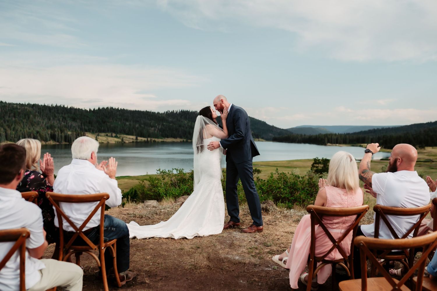 A wedding kiss during a scenic outdoor ceremony with mountains and lake in the background.