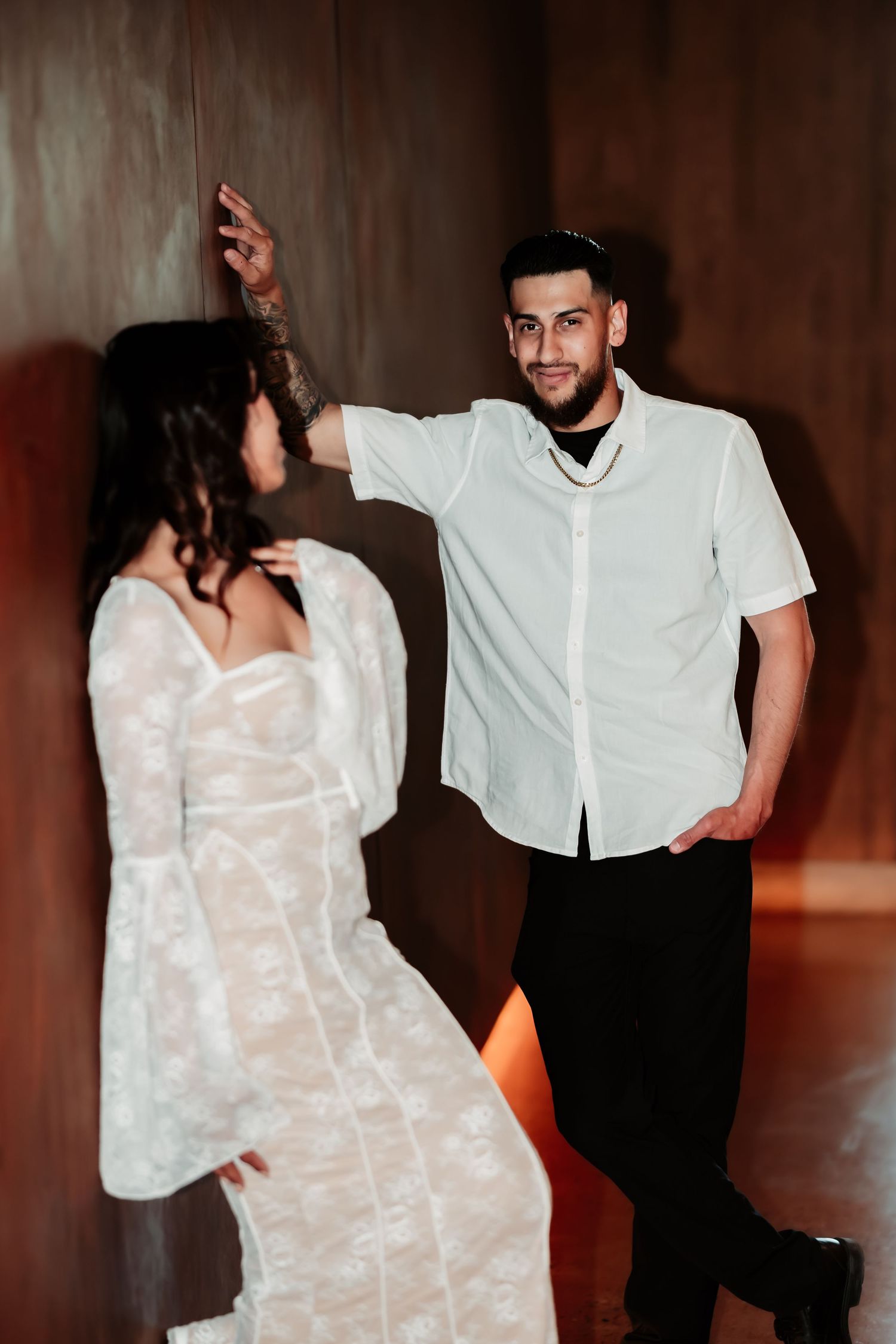 A romantic couple in white outfits dancing together against a moody dark wall with dramatic lighting.