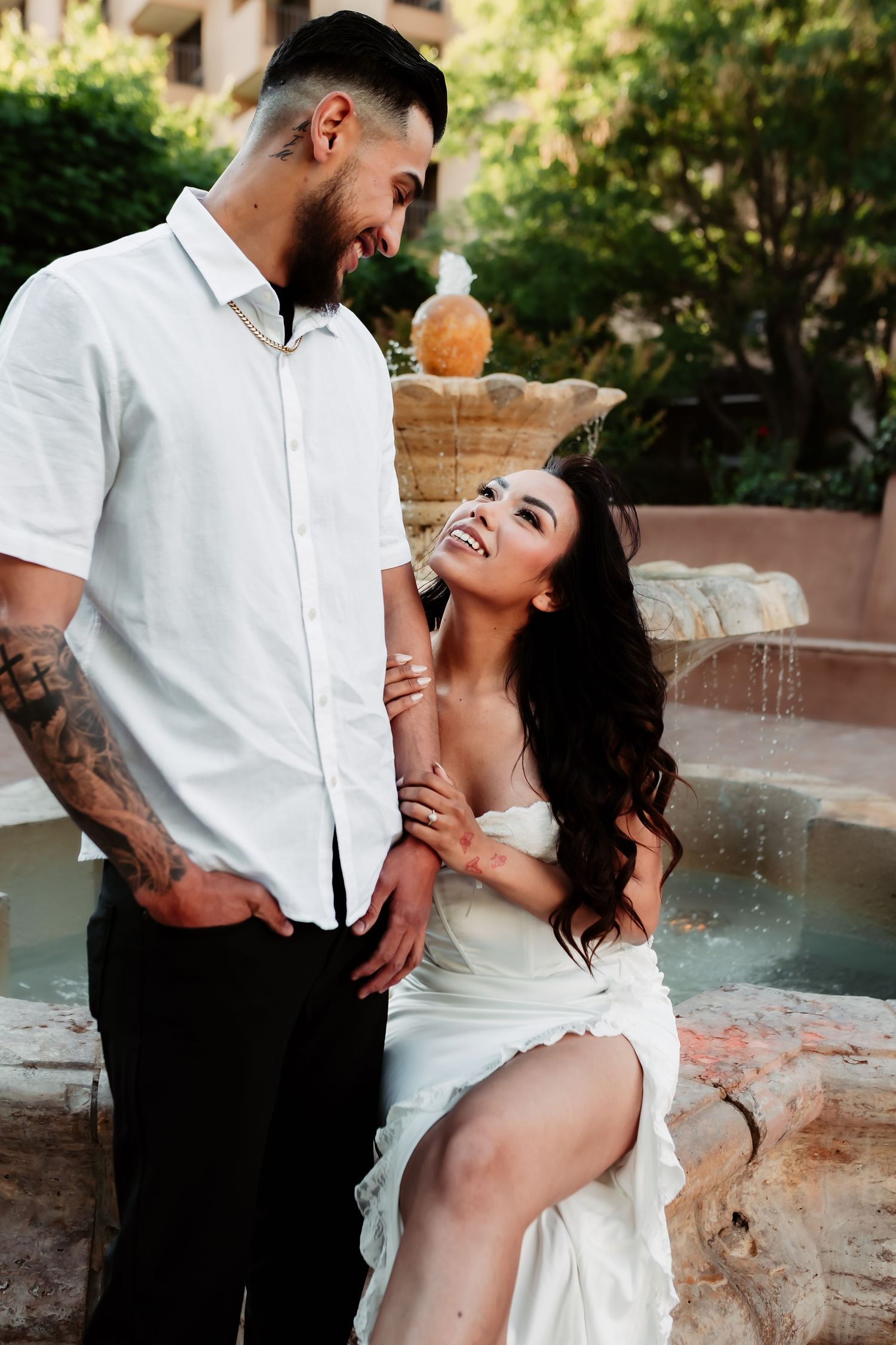 A couple poses romantically by a fountain, with one standing and one seated on the fountain edge.