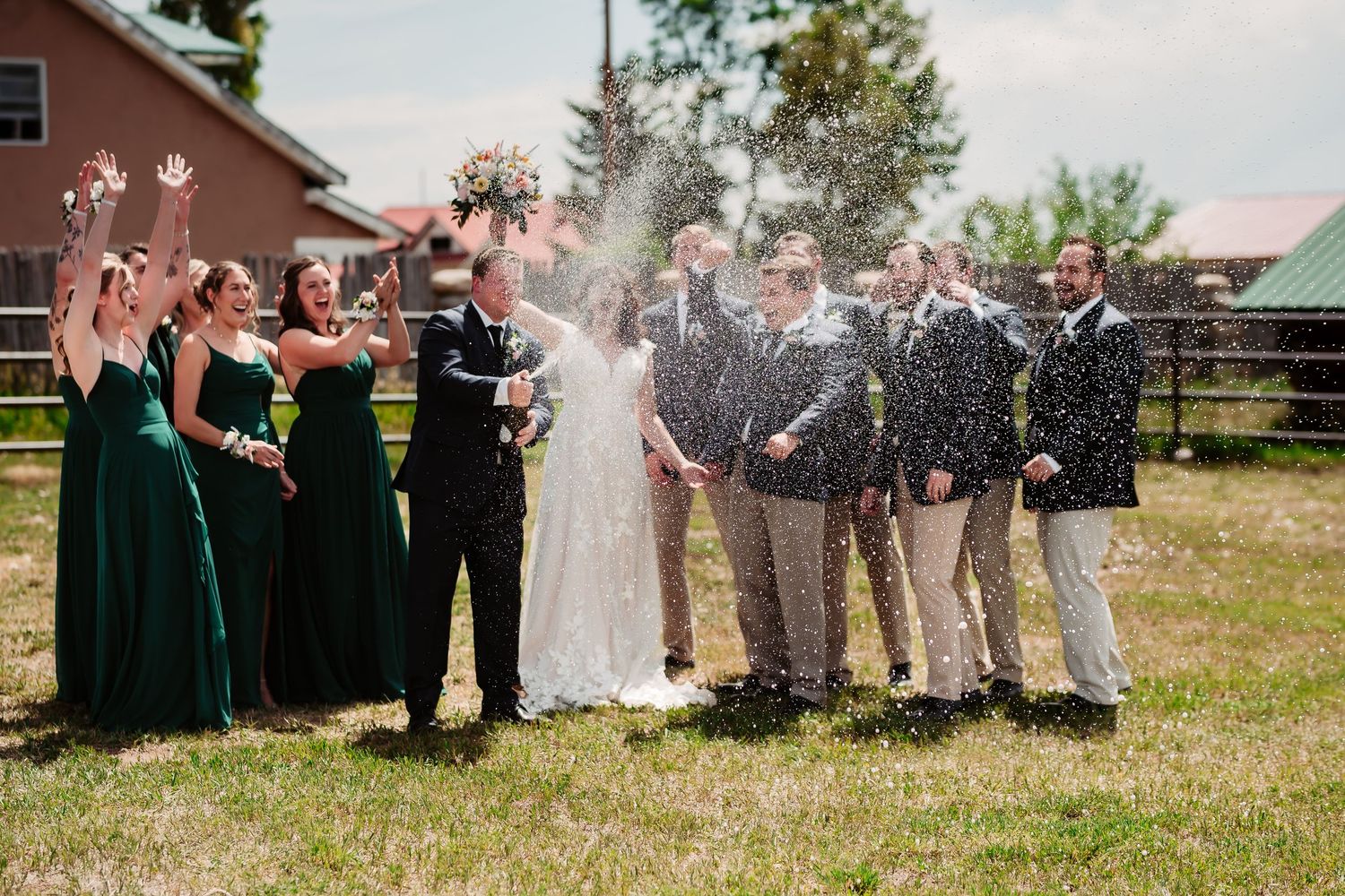 Wedding party in emerald green dresses and gray suits celebrate outdoors with champagne spray during a festive moment.