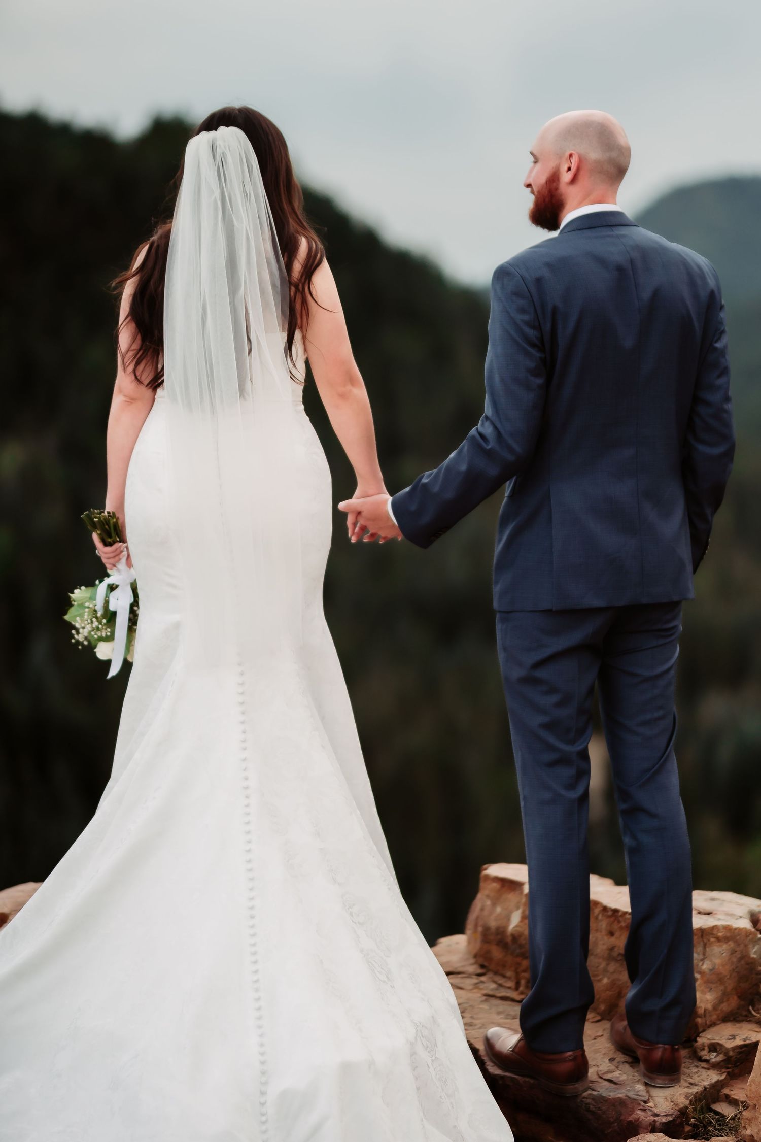 Couple in wedding attire hold hands while standing on rocky overlook with mountain views.