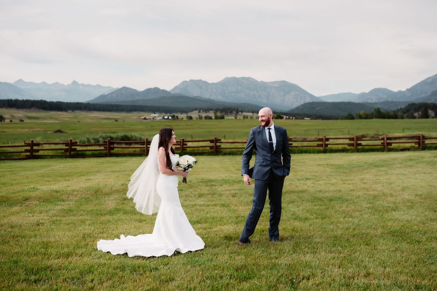 Wedding couple posing in scenic mountain meadow with fence and peaks in background.